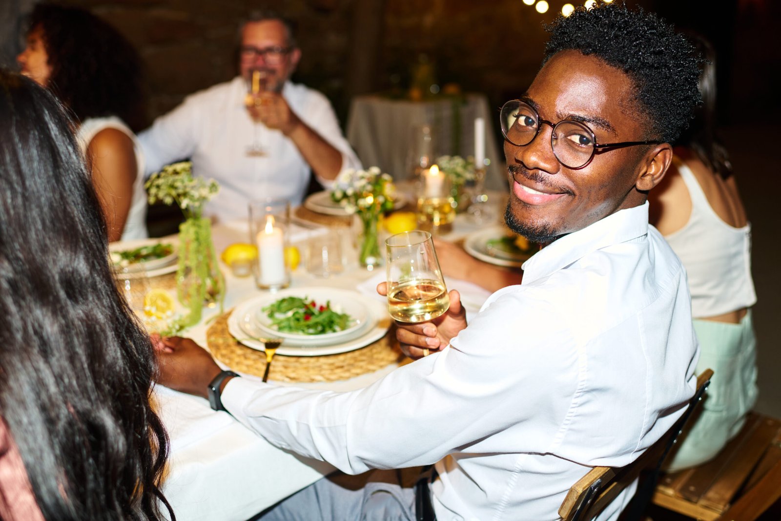 Happy young African American man with flute of champagne sitting by served table and cheering up with friends at wedding feast